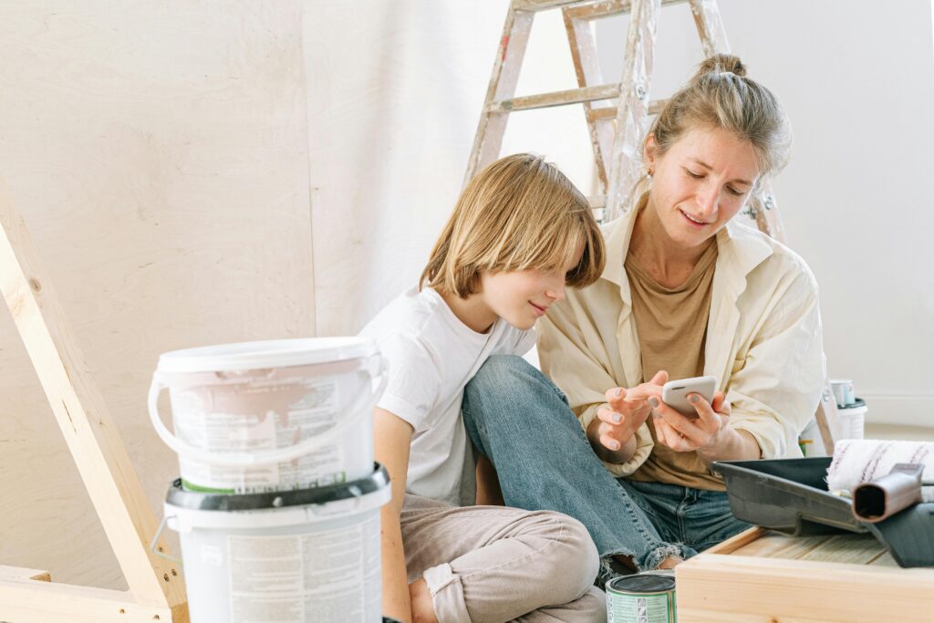 A mother and son share a moment painting a room while looking at a smartphone.